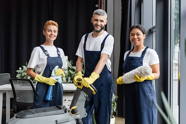 Three janitors in uniforms and gloves stand with cleaning equipment in an office setting, smiling at the camera.