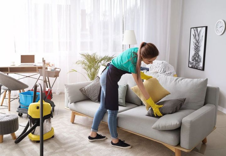 Person wearing gloves and apron arranges cushions on a gray sofa in a tidy living room with cleaning supplies nearby.