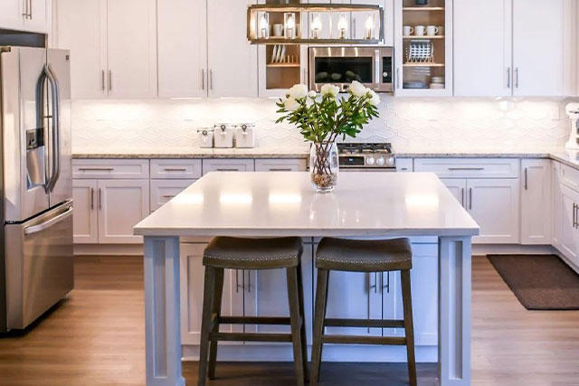 Modern kitchen with white cabinets, a central island with two stools, stainless steel appliances, and vase of flowers.